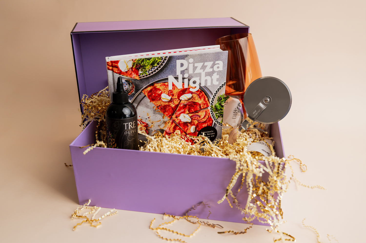 Purple gift box with pizza-themed items including a book, bottle, and pizza cutter on a beige background.