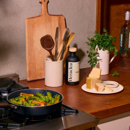Cooking scene with pan on stove, cutting board, utensils, and food items on a wooden counter.