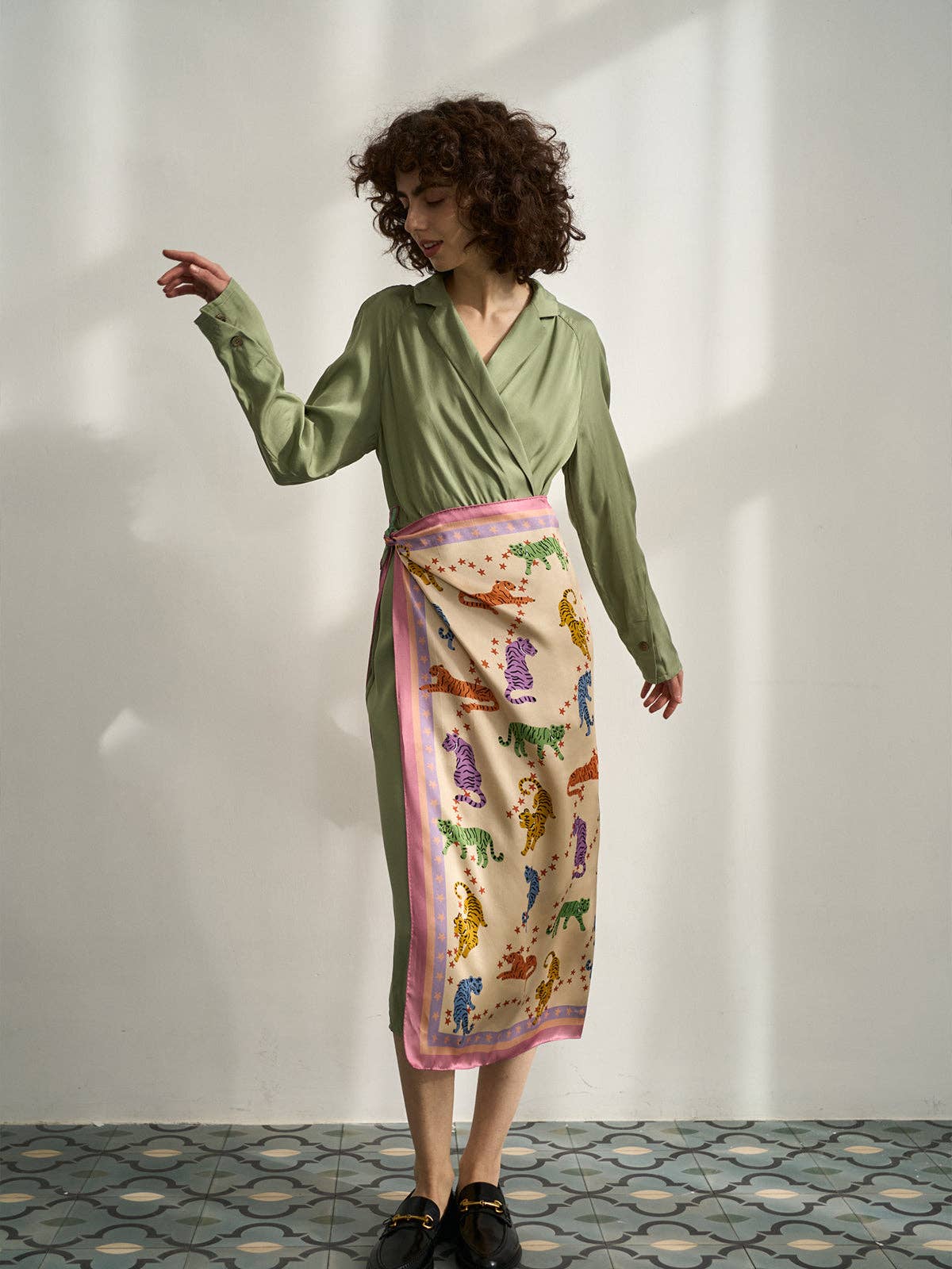 Woman wearing a green dress with a colorful scarf around her waist against a white wall.